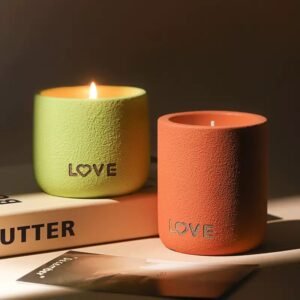Three candle cups in cream, gray, and green arranged on a wooden tray with a burning candle inside one.